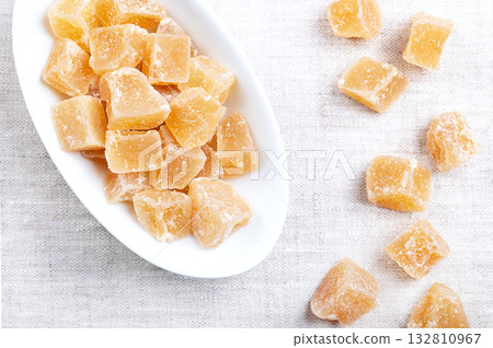 Candied ginger, crystallized ginger roots in a white bowl on linen. Rhizomes of Zingiber officinale, cut into block shaped chunks, boiled, and preserved with sugar. A sweet snack for the cold season. Candied ginger, crystallized ginger roots in a white bowl on linen. Rhizomes of Zingiber officinale, cut into block shaped chunks, boiled, and preserved with sugar. A sweet snack for the cold season. 132810967
