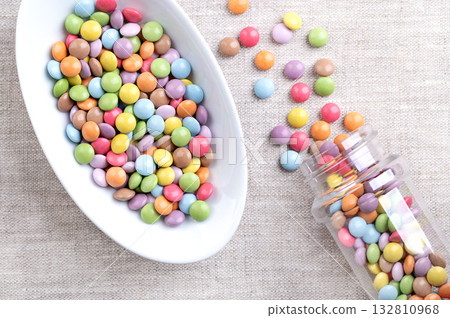 Candy coated mini chocolate lentils, in a white bowl, on linen. Small, sugar-coated chocolate dragees, with multi colored candy shell. Tiny dragee chocolate confectionery, used as colorful sprinkles. 132810968