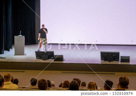 Speaker on stage delivering presentation to audience in conference hall with large screen Speaker on stage delivering presentation to audience in conference hall with large screen 132810974