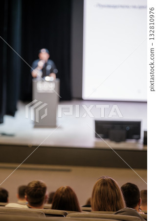 Speaker on stage addressing audience at conference with blurred projection screen 132810976