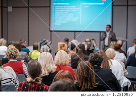 Business conference audience listens to presenter in modern conference room with projector screen 132810980