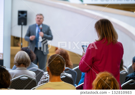 Business conference audience listens to speaker as woman in red records presentation on phone Business conference audience listens to speaker as woman in red records presentation on phone 132810983