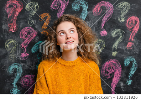 Young woman in yellow sweater looking up in front of blackboard with colorful question marks 132811926