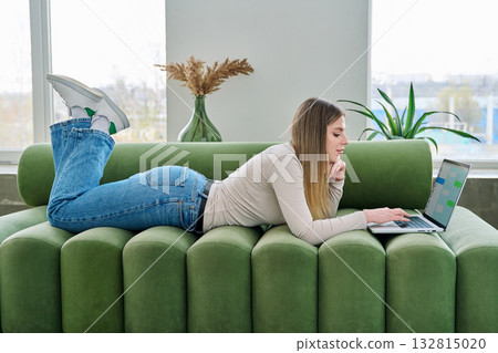 Young woman lying on sofa at home using laptop, texting in messengers Young woman lying on sofa at home using laptop, texting in messengers 132815020