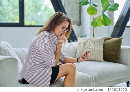 Sad serious middle aged woman with smartphone sitting on sofa Sad serious middle aged woman with smartphone sitting on sofa 132815050