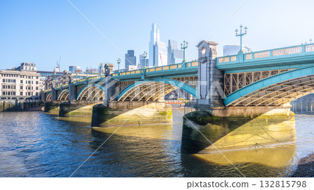 Bright sunlight reflects off the Thames River as Southwalk Bridge stands proudly in London. The bridge connects the bustling cityscape with scenic views along the riverbank. 132815798