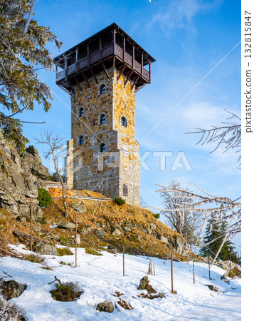 A historic lookout tower stands on Wysoki Kamien in the Jizera Mountains of Poland, surrounded by snow-covered ground, tall trees, and a clear blue sky, inviting visitors year-round. A historic lookout tower stands on Wysoki Kamien in the Jizera Mountains of Poland, surrounded by snow-covered ground, tall trees, and a clear blue sky, inviting visitors year-round. 132815847