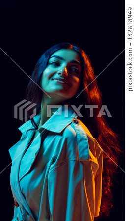 Confident woman in trench coat under vivid cinematic light looking upward with soft smile 132818949