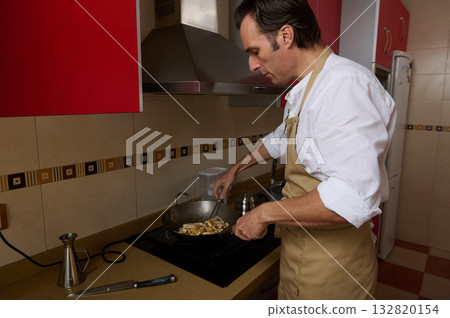Man Cooking In Kitchen With Apron At Stove, Preparing Food In Wok, Home Cooking Scene Man Cooking In Kitchen With Apron At Stove, Preparing Food In Wok, Home Cooking Scene 132820154
