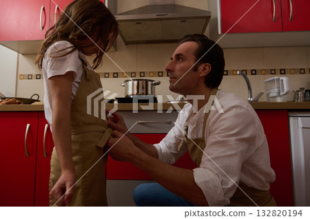 Father Helps Daughter In Cozy Kitchen, Cooking Soup Together With Aprons On 132820194