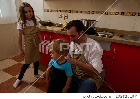 Father Cooking Soup With His Kids In Modern Kitchen, Helping And Watching Them Father Cooking Soup With His Kids In Modern Kitchen, Helping And Watching Them 132820197