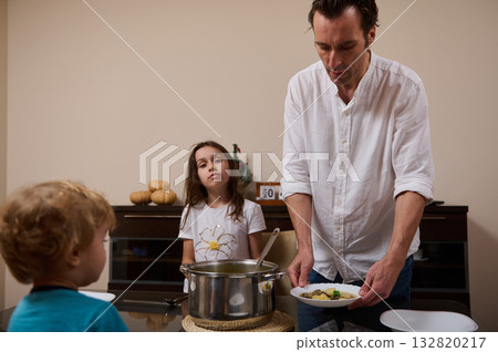 Father Cooking Soup and Serving Dinner to His Kids in a Cozy Kitchen Father Cooking Soup and Serving Dinner to His Kids in a Cozy Kitchen 132820217