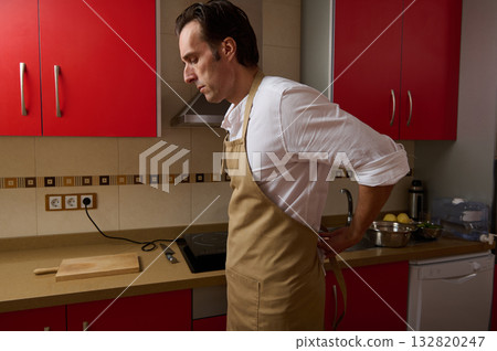 Focused Male Chef In Beige Apron Preparing Dinner In Modern Kitchen With Red Cabinets 132820247