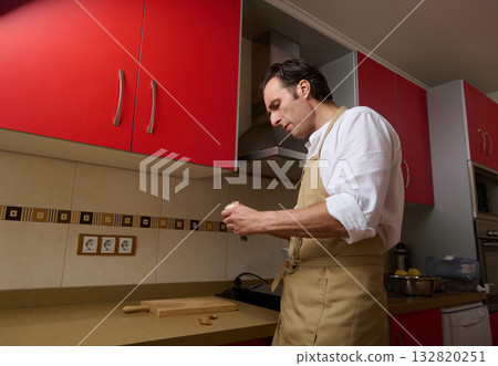 Man In Beige Apron Preparing Dinner In A Modern Kitchen With Red Cabinets Man In Beige Apron Preparing Dinner In A Modern Kitchen With Red Cabinets 132820251