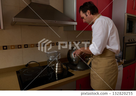 Man In Apron Cooking In Modern Kitchen, Focused On Washing A Pot By The Stove 132820255