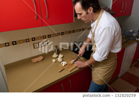 Chef in Beige Apron Chopping Vegetables on Kitchen Counter in Red-Cabinet Kitchen, Modern Home Culinary Scene 132820259