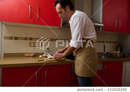 Man Cooking In Modern Kitchen Wearing Apron While Daughter Helps Prepare Dinner Man Cooking In Modern Kitchen Wearing Apron While Daughter Helps Prepare Dinner 132820260