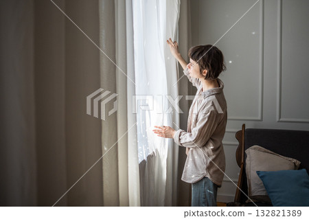 Young relaxed woman standing beside window in cozy living room and opening white curtains in morning Young relaxed woman standing beside window in cozy living room and opening white curtains in morning 132821389