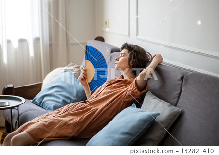 Tired woman lying resting on sofa waving hand paper fan, suffering from summer heat, annoyed face. Tired woman lying resting on sofa waving hand paper fan, suffering from summer heat, annoyed face. 132821410