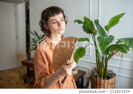 Woman standing near window in living room waving hand fan for cooling, suffering from overheating. 132821434