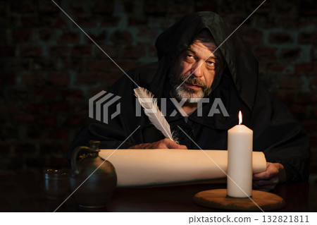 A man in a dark hoodie works on a manuscript in a mysterious setting. 132821811