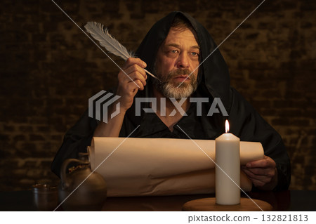 A hooded man with a quill in his hand is immersed in work on a manuscript. 132821813