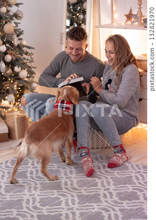 Couple opens Christmas gift with their dog on a joyful Christmas morning. The scene expresses love, warmth and the magic of the holiday season. 132821970