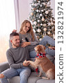 Smiling couple relaxes with their dog by a Christmas tree. The scene reflects love warmth and the joyful spirit of the holiday season. 132821974