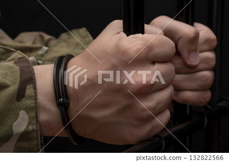 Handcuffed soldier clutching the bars with his hands against a black background. Concept: trial of a soldier, desertion in the army, prisoner of war. Handcuffed soldier clutching the bars with his hands against a black background. Concept: trial of a soldier, desertion in the army, prisoner of war. 132822566