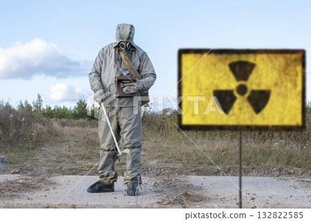 A military man in chemical protection and a gas mask measures the radiation level with a dosimeter, a radiation hazard sign, a concept: radiation leakage, the threat of infection. 132822585