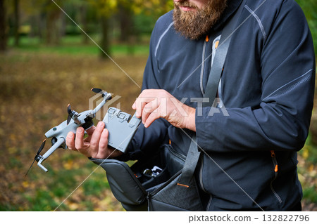 A man with a bushy beard prepares his drone for flight in a peaceful park filled with trees. He carefully adjusts the device on a calm, sunny day. A man with a bushy beard prepares his drone for flight in a peaceful park filled with trees. He carefully adjusts the device on a calm, sunny day. 132822796