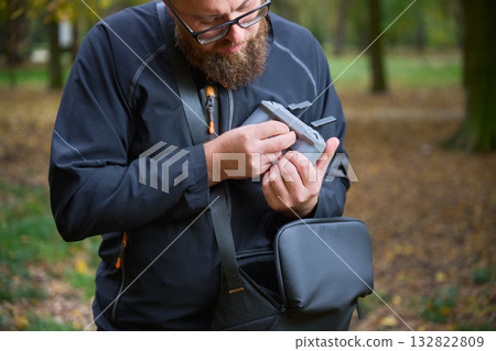 A person examines a camera accessory closely while standing in a beautiful autumn park. Surrounding yellow and orange leaves create a peaceful atmosphere. 132822809