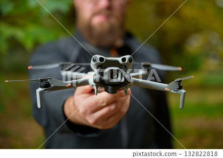 An individual proudly displays a cutting-edge drone, its sleek design gleaming. Surrounding trees exhibit vibrant fall colors, creating a perfect backdrop for this tech moment. 132822818