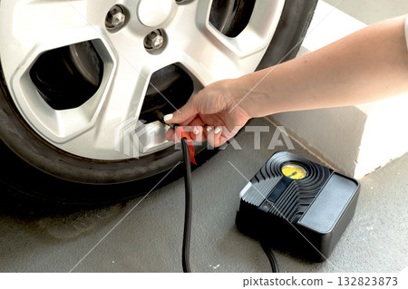 Close-up of woman inflating car tire using portable electric air compressor. DIY vehicle maintenance at home or garage. 132823873