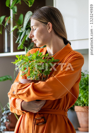 Relaxed smiling woman holding pot with homegrown peppermint in hands, enjoys indoor gardening hobby. Relaxed smiling woman holding pot with homegrown peppermint in hands, enjoys indoor gardening hobby. 132824989