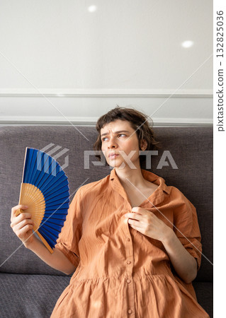 Tired female lying on couch waving hand fan for cooling, suffering from summer heat. exhausted face. Tired female lying on couch waving hand fan for cooling, suffering from summer heat. exhausted face. 132825036