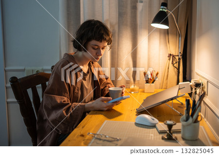 Tired woman scrolling smartphone late night. Drinking evening tea after laptop work at home 132825045