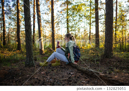 Female hiker taking break after walk in forest and forage, sitting on log drinking water from bottle 132825051
