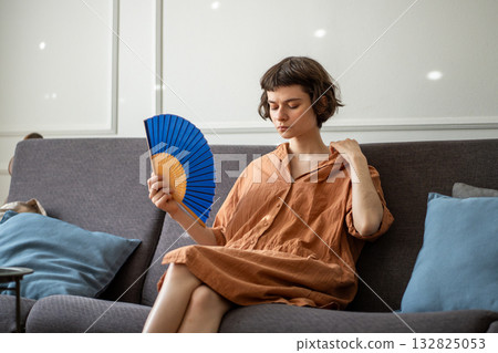 Woman sitting on sofa and cooling, waving hand paper fan, suffering from summer heat, annoyed face. 132825053