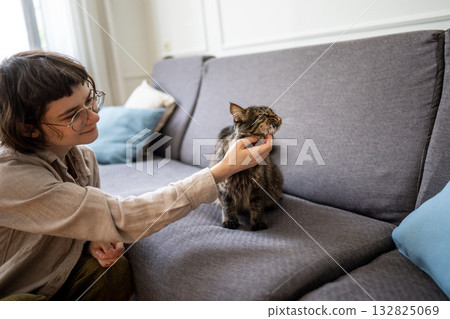 Young cheerful woman pet owner spending time at home with cat, sitting beside sofa petting her kitty Young cheerful woman pet owner spending time at home with cat, sitting beside sofa petting her kitty 132825069