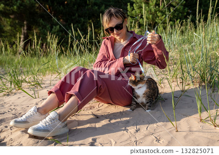 Woman sitting on beach coast with her pet, worried domestic cat on leash exploring wild nature. Woman sitting on beach coast with her pet, worried domestic cat on leash exploring wild nature. 132825071