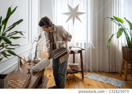 Woman housekeeper tidying in apartment room, wiping dust with feather duster, cleaning living space. Woman housekeeper tidying in apartment room, wiping dust with feather duster, cleaning living space. 132825073