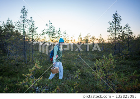Woman hiker with basket in hand walking out of autumn forest at twilight, returns home after forage. 132825085