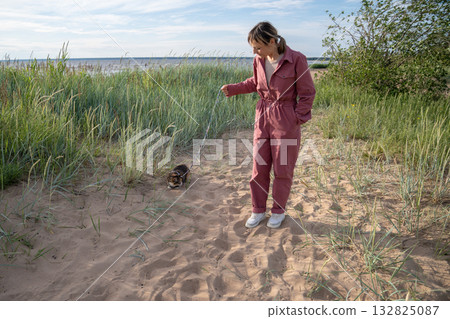 Young woman walking on beach together with her pet, guiding domestic cat on leash exploring nature. 132825087