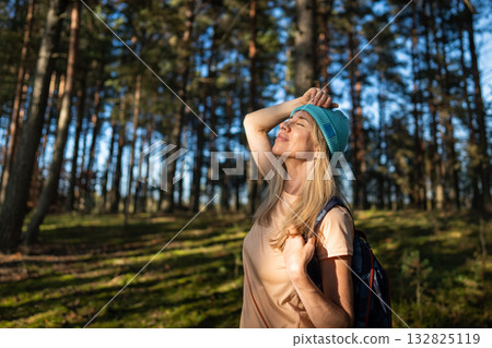 Joyful female traveler blinking enjoying sunlight while walking through woodland with backpack 132825119