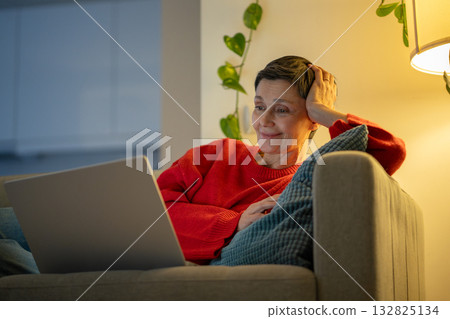 Satisfied senior woman reclines comfortably browsing laptop on sofa. Cozy downtime, home comfort 132825134