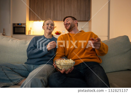 Smiling boyfriend and girlfriend with popcorn watching TV and having fun at home, laughing couple 132825137