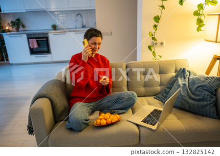 Laughing woman chats on phone while peeling mandarins and using laptop on sofa. Happy retirement Laughing woman chats on phone while peeling mandarins and using laptop on sofa. Happy retirement 132825142