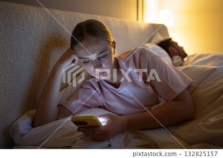 Exhausted woman using smartphone in bed at night struggling with sleep due to social media overuse 132825157