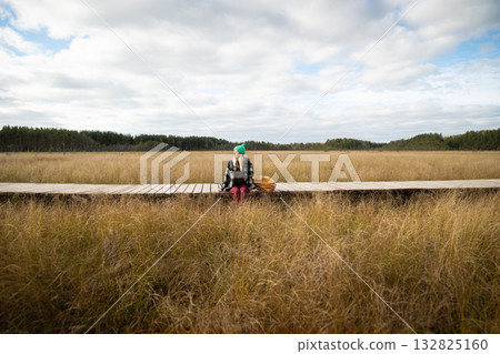 Relaxed woman sitting on wooden trail on autumn peat bog and resting surrounded by dry sedge grass.  132825160